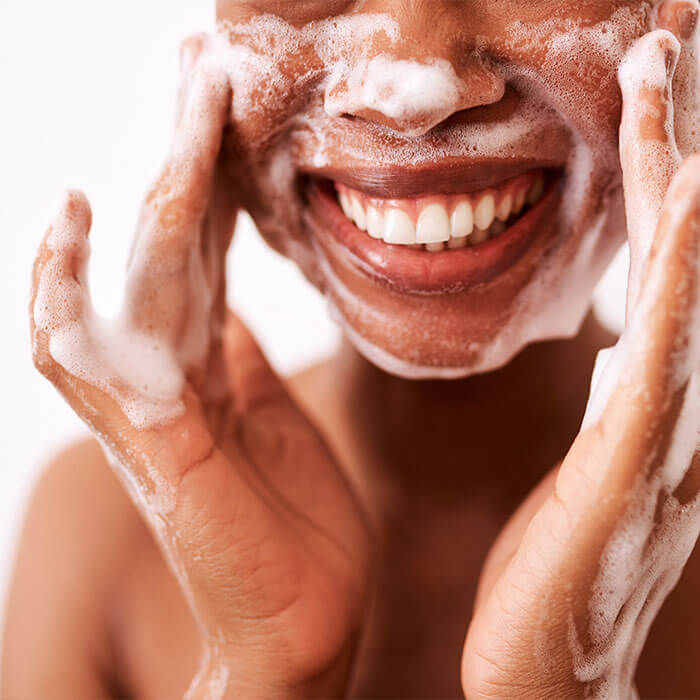 An image of a woman of color, smiling at the camera as she washes her face