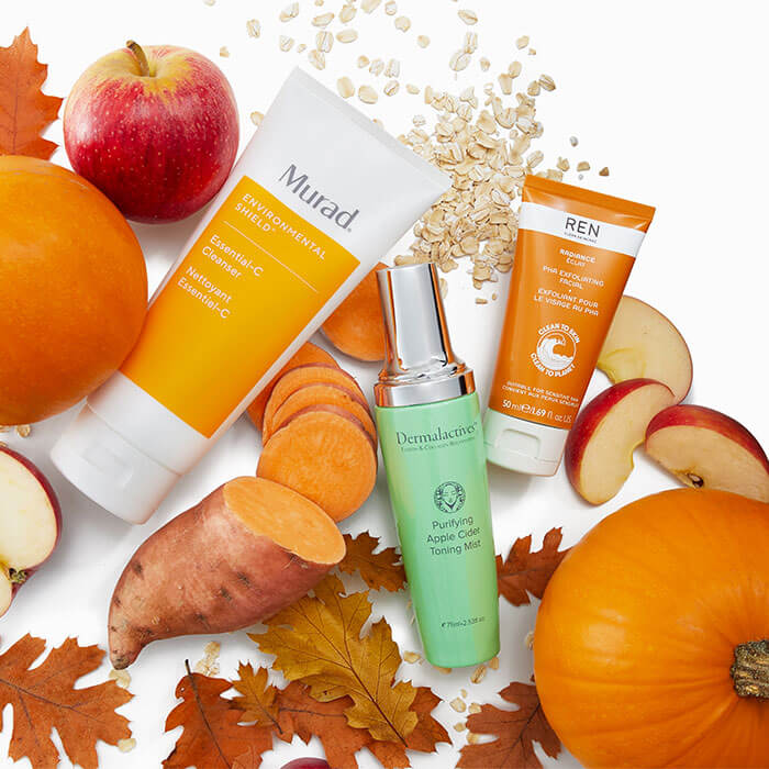 Various skincare products scattered across white background surrounded by fall leaves, apples, pumplins, and sweet potato