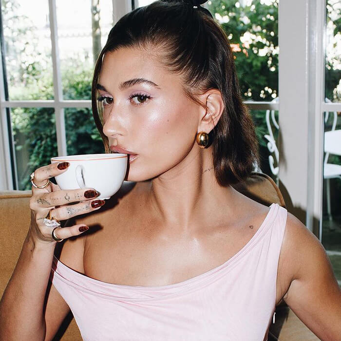 An image of Hailey Bieber wearing a pink off-shoulder casual dress accessorized with a golden earring, while enjoying a cup of coffee