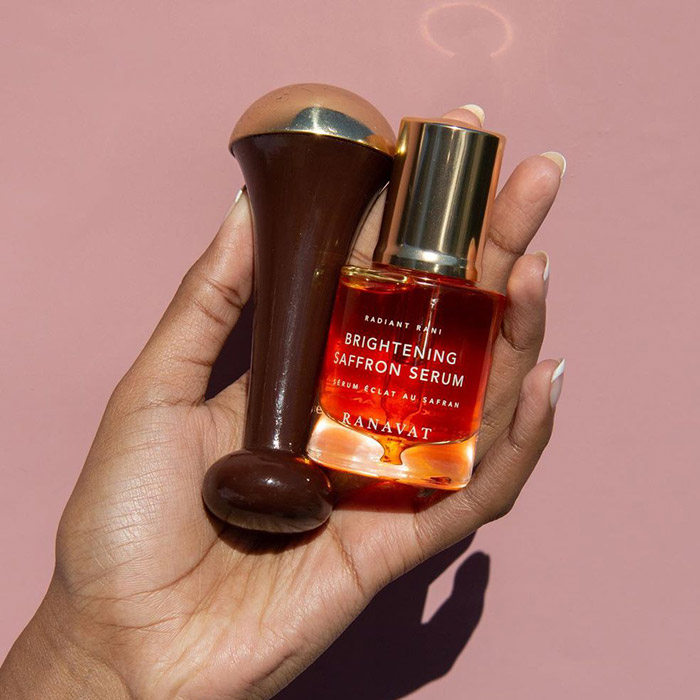 A photo of a woman's hand holding Kansa Wand and Brightening Saffron Serum on a rose gold background