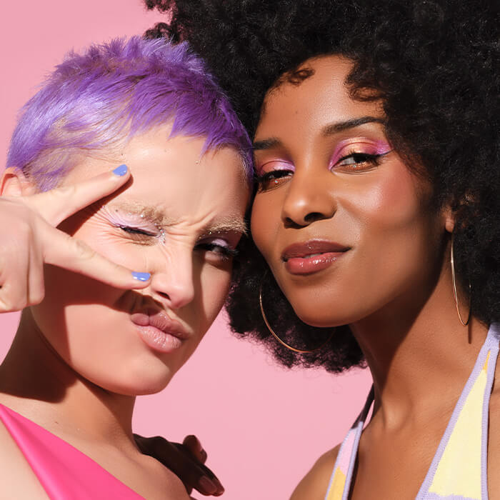 Two young models rocking purple hair and colorful eyeshadow makeup looks posing in front of pink background