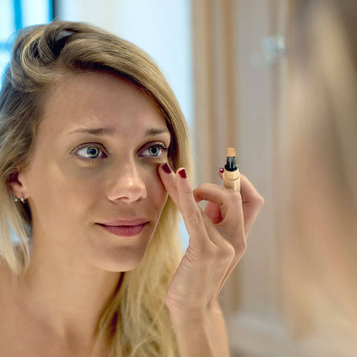 A woman using a concealer stick to apply makeup under her eyes, demonstrating techniques for hiding scars with makeup
