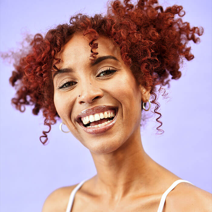 An image of a woman with color with a curly red hair styled in a two-piece messy bun wearing a bright blue spaghetti top while grinning at the camera