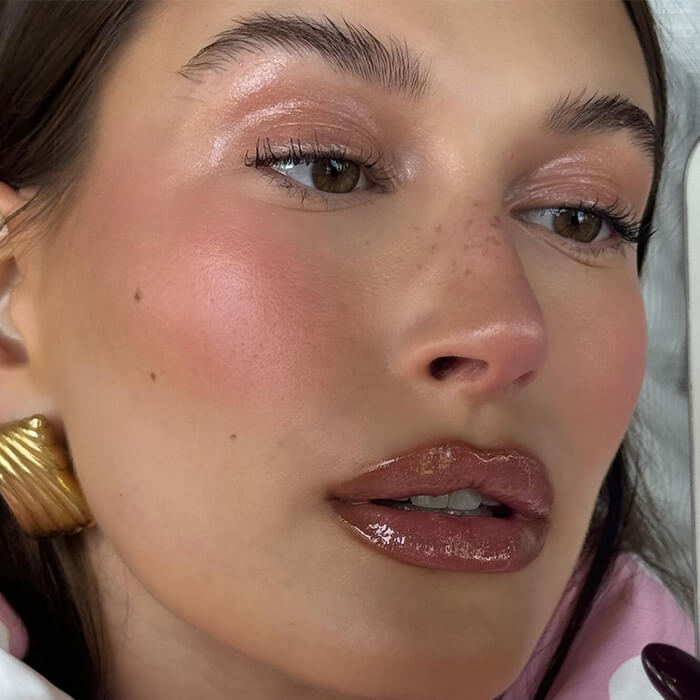An image of Hailey Bieber sporting a natural makeup look with pink blush, eyeshadow, and lip gloss, complemented by a gold earring
