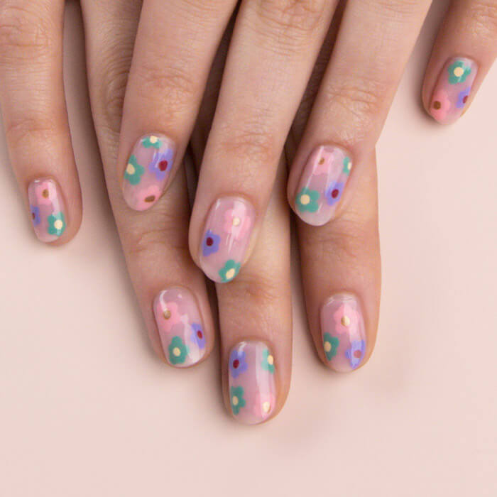 Close-up of model's hands with a purple, pink, and green flower nail art mani on light peach background