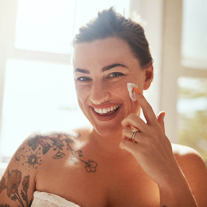 Close-up image of a woman with a shoulder tattoo smiling while applying face cream on her cheek